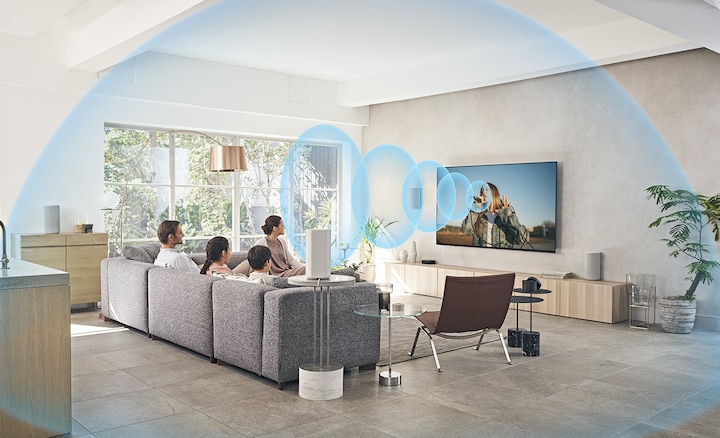 Familia en un sofá viendo el TV con el sistema de Home Theatre HT-A9 en un mueble de madera con ondas de sonido emanando de la pantalla.