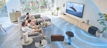 Familia en un sofá viendo el TV con el sistema de Home Theatre HT-A9 en un mueble de madera con ondas de sonido emanando del subwoofer.