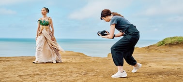 Imagen de uso en la que se muestra a una mujer con una cámara manual que hace capturas a otra mujer con un traje de época y la costa de fondo